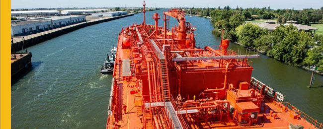 Photograph of a bright organge, large cargo ship with a tug boat at it's side docking on a river.