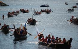 Photograph of ceremonial canoes on the water, depicting the gathering of Salish Coast people who travel through the waterways of the Salish Sea to annually to celebrate their connection to salmon, water and each other.