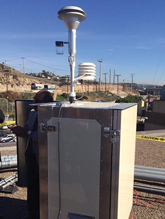 Air monitor mounted on roof with cars and hillside in background. Five foot tall silver box with an air intake pipe with rain shield emerges from top of box pointing up to catch the air flow.
