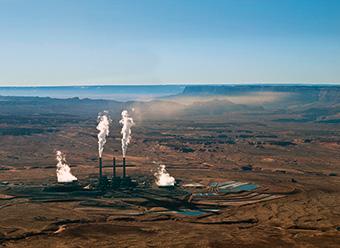 Aerial view of desert scene with mesas in distance. NGS in forground with white steam emanating from smokestacks and cooling towers. Light brown haze drifts from top of smokestack plume to the right and back towards mesas. Photo: Ted Grussing