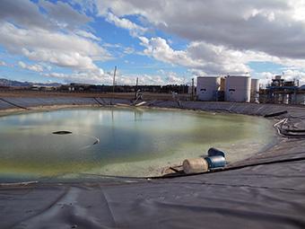 Black plastic sheeting material lines a depression filled with green liquid with 2 plastic barrels at waters edge, white metal storage tanks in background.