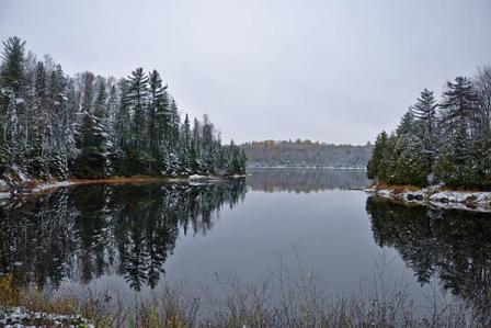 Conifers dusted with snow are reflected in Deer Lake on a gray day