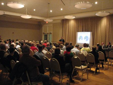 This image shows a public meeting that took place in Williamsburg, Virginia. The image shows the backs of the audience, the speaker at a podium and slides projected onto a screen next to the speaker.