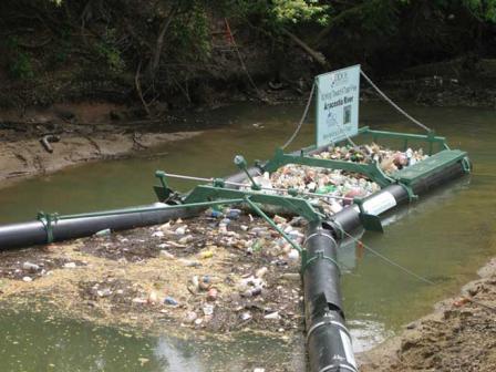 Inflatable booms channel floating trash onto what appears to be a raft with open arches allowing trash in. Trash leel appears higher than water level.