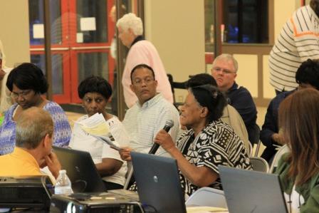 People at a meeting listening to a woman speaking, holding a microphone.