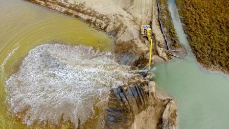 Aerial view looking down on an earthmover digging a breach in the levee at sears point. Water is pouring through the breach from right to left and flooding a pool.